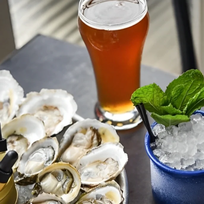 A tray of opened oysters with sauce droppers next to a glass of amber beer and a blue cup with crushed ice and mint leaves.