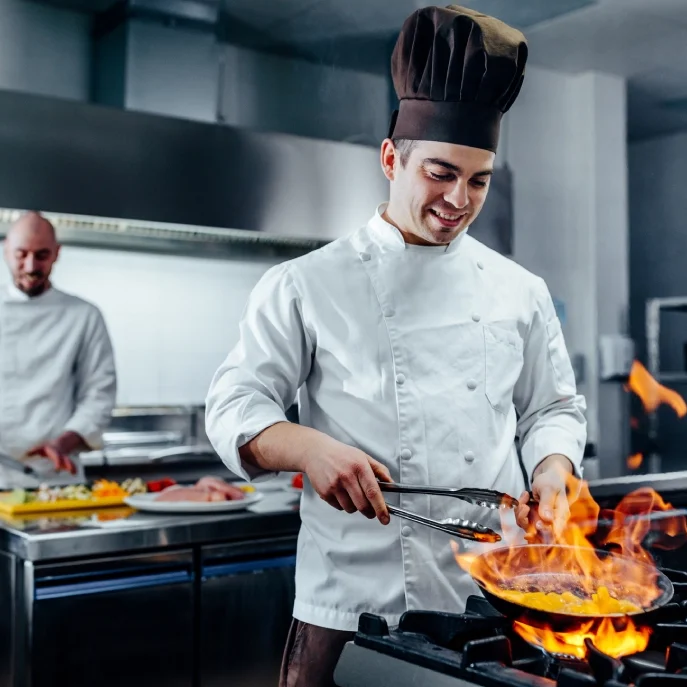 Chef in a white uniform and black hat flambés food in a pan over a stove, while another chef works in the background, slicing ingredients on a cutting board in a professional kitchen.